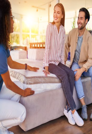 African American saleswoman helps customers choose an orthopedic mattress in a store. A young couple is buying a mattress and bed and various bedding accessories.